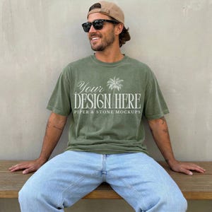 Puede incluir: Una camiseta de manga corta color verde oliva con texto en blanco que dice Your DESIGN HERE y PIPER & STONE MOCKUPS, junto con un gráfico de palmera blanca. La camiseta se combina con jeans azul claro, una gorra de béisbol beige y gafas de sol.