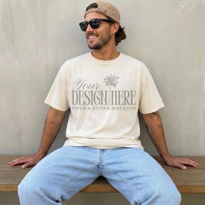 May include: A cream-coloured short-sleeve t-shirt featuring the grey text "YOUR DESIGN HERE" and "PIPER & STONE MOCKUPS" with a palm tree graphic centred on the chest. The shirt is paired with light blue denim jeans, a tan baseball cap, and sunglasses.