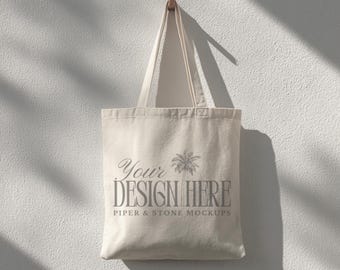 May include: A cream-colored canvas tote bag with two shoulder straps. The front of the bag features a gray graphic print of a palm tree and the text "YOUR DESIGN HERE" followed by the smaller text "PIPER & STONE MOCKUPS" in a serif font.