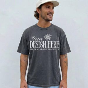 Può includere: Una t-shirt a maniche corte color grigio antracite con una grafica bianca di una palma e il testo "YOUR DESIGN HERE" seguito da "PIPER & STONE MOCKUPS" in un carattere serif. La maglietta presenta un girocollo e una vestibilità ampia.
