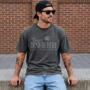 May include: A charcoal gray short-sleeve t-shirt featuring a palm tree graphic and the text "YOUR DESIGN HERE" and "PIPER & STONE MOCKUPS" in a light gray font. The shirt has a crew neckline and a relaxed fit, worn with light blue denim jeans.