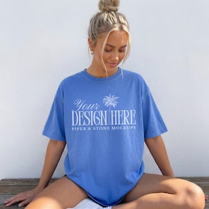 Può includere: Una t-shirt a maniche corte color blu pervinca con testo serigrafato in bianco che recita Your DESIGN HERE e PIPER & STONE MOCKUPS, accompagnata da un'illustrazione grafica di una palma centrata sopra il testo.