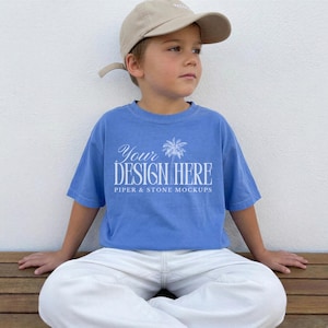 May include: A light blue short-sleeve t-shirt featuring a white graphic of a palm tree and the text "YOUR DESIGN HERE" followed by "PIPER & STONE MOCKUPS" in a white serif font, paired with a beige baseball cap and white pants.