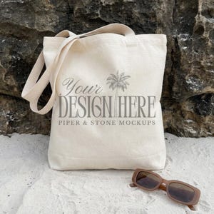 Natural cotton canvas tote bag mockup featuring the OAD113 Liberty Bags style displayed on soft beach sand against a coastal rock backdrop with sunglasses, creating a relaxed lifestyle aesthetic perfect for POD and print on demand sellers showcasing shopping bag designs as a digital download