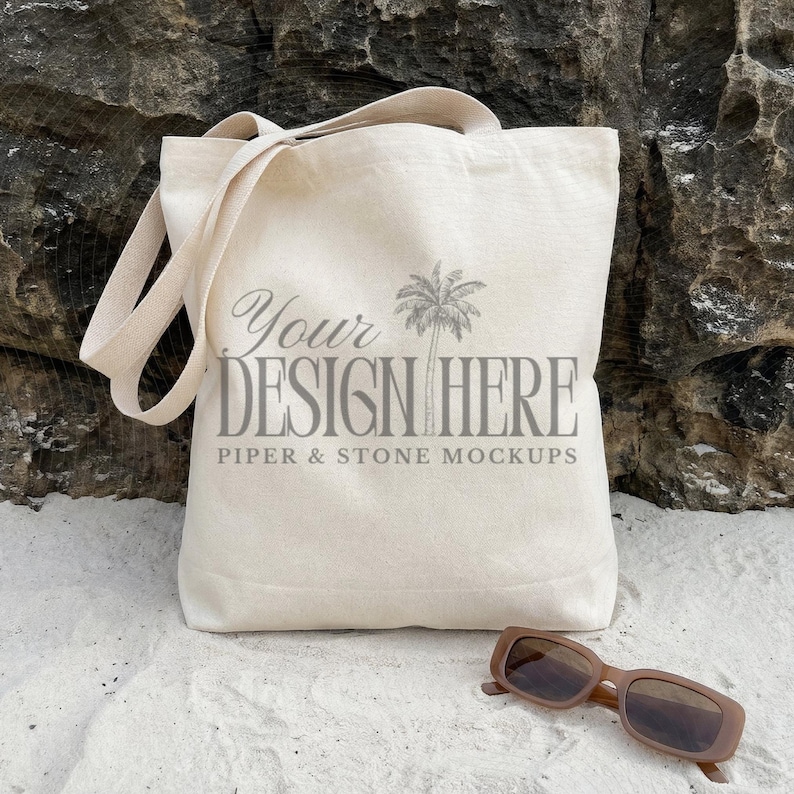 Natural cotton canvas tote bag mockup featuring the OAD113 Liberty Bags style displayed on soft beach sand against a coastal rock backdrop with sunglasses, creating a relaxed lifestyle aesthetic perfect for POD and print on demand sellers showcasing shopping bag designs as a digital download