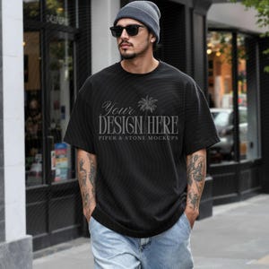 Puede incluir: Una camiseta negra de manga corta con un gráfico de palmera y el texto "YOUR DESIGN HERE" seguido de "PIPER & STONE MOCKUPS" en una fuente serif. La camiseta tiene un corte holgado y se lleva con vaqueros azul claro y un gorro gris.