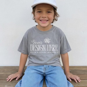 May include: A grey short-sleeve t-shirt featuring white text that reads YOUR DESIGN HERE with a palm tree graphic and the smaller text PIPER & STONE MOCKUPS below it. The shirt is worn with blue denim jeans and a white baseball cap.