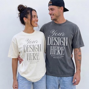 May include: A cream-colored t-shirt and a charcoal gray t-shirt, both featuring the text "Your DESIGN HERE" and "PIPER & STONE MOCKUPS" printed in a serif font on the front. The shirts have short sleeves and a crew neckline, worn with blue jeans.