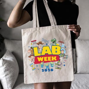 May include: Beige canvas tote bag featuring a colorful graphic with the text "LAB WEEK 2026" in yellow and blue. The design includes cartoon characters and science-themed illustrations. The bag has two beige handles.