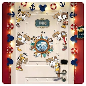 May include: A white door decorated with Disney-themed cutouts, including Mickey Mouse, Donald Duck, and Goofy, holding signs that say "Name". A central nautical wheel design features the word "Alaska". Decorative lights and nautical accents complete the theme.