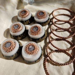 May include: A collection of handcrafted resin art pieces. The image features seven round, layered resin objects with copper wire accents and a set of copper-colored twisted metal bracelets. The resin pieces have a white, gray, and gold color scheme.