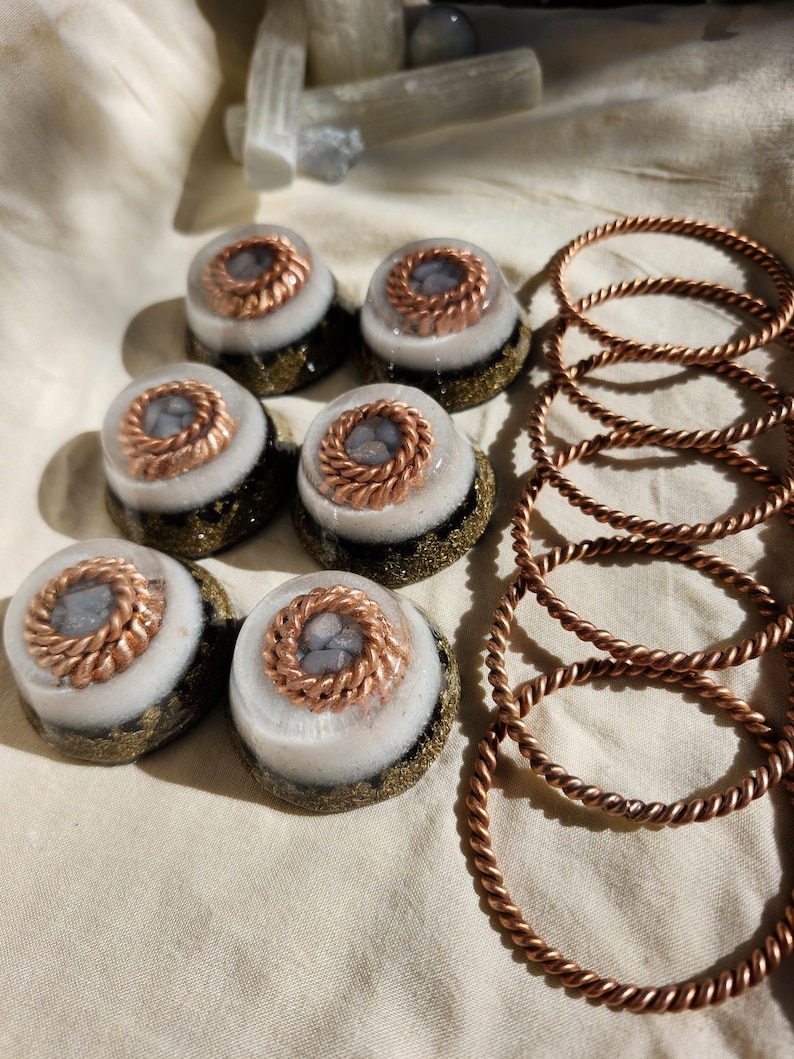May include: A selection of handcrafted resin art pieces. The image shows seven round, layered resin objects with copper wire accents and a set of copper-coloured twisted metal bracelets. The resin pieces have a white, grey, and gold colour scheme.