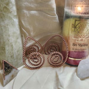 May include: A pair of copper-colored earrings featuring a twisted wire design that forms a double spiral shape. The earrings are positioned next to a tall candle with a Sri Yantra symbol and text, a pyramid-shaped object, and a small white stone.