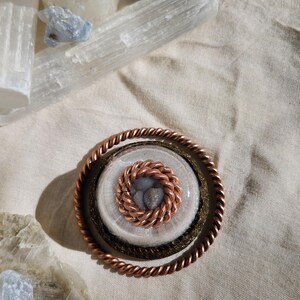 May include: A copper pendant with a twisted rope design around the outer edge. The centre features a smaller, woven copper circle surrounding a cluster of small, grey stones. The pendant rests on a beige fabric, with crystals and stones in the background.