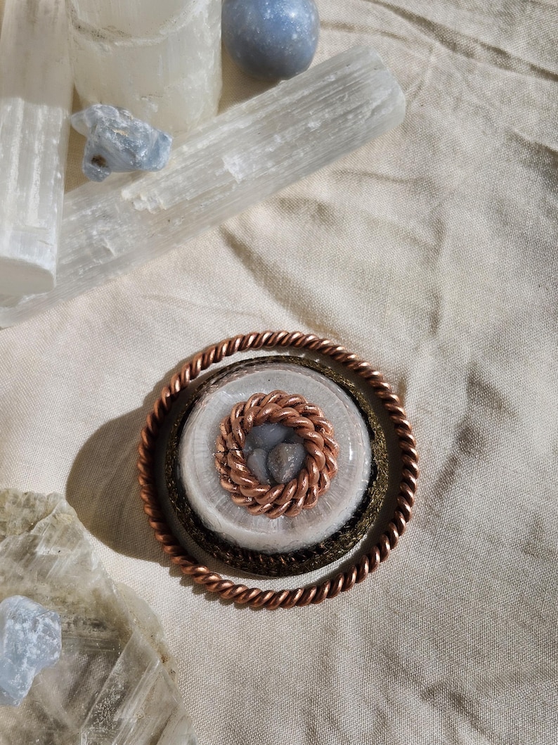 May include: A copper pendant with a twisted rope design around the outer edge. The centre features a smaller, woven copper circle surrounding a cluster of small, grey stones. The pendant rests on a beige fabric, with crystals and stones in the background.