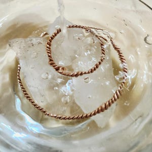 May include: A copper-colored twisted wire ring and a matching twisted wire bracelet resting on clear ice cubes submerged in water. The metal has a warm, metallic sheen, and the twisted texture creates a continuous spiral pattern throughout.