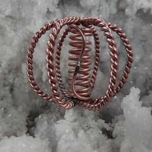 May include: A copper-colored metal sculpture with a spiral center and three twisted rings. The sculpture is set against a white, textured background, creating a contrast of textures and colors. The design is intricate and geometric.