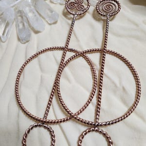 May include: Two copper-colored metal hair pins featuring a twisted wire design. Each pin has a long straight stem topped with a circular decorative element, one featuring a triskelion pattern and the other a spiral, with a large ring at the base.