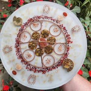 May include: A circular resin disc featuring a central copper wire grid, embedded with tiger's eye stones, small red mineral chips, two spiral ammonite fossils, and various copper wire coils arranged in a geometric pattern on a translucent base.