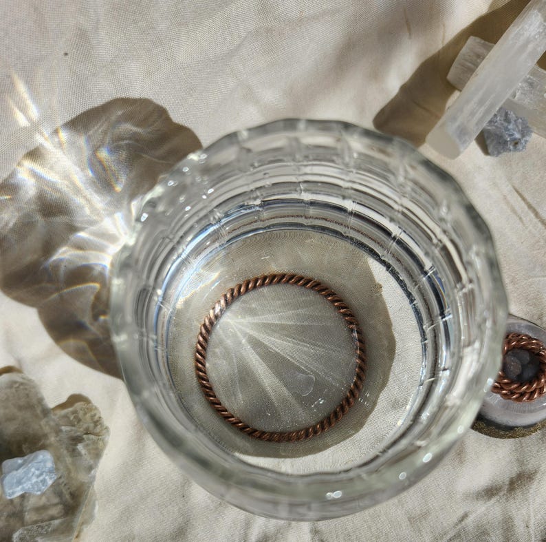 May include: A clear glass with a copper-coloured ring inside, filled with water. The ring has a twisted rope design. The glass sits on a beige surface, with light reflecting off the water. Crystals are visible in the background.