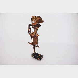 Puede incluir: Una marioneta tradicional indonesia Wayang Kulit. La marioneta está hecha de cuero tallado y pintado intrincadamente, con un diseño detallado en colores marrón, dorado y negro. Se encuentra sobre una pequeña base negra con detalles dorados, mostrando la artesanía.