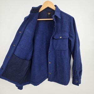 May include: A navy blue wool-blend shirt jacket with a button-down front, two chest pockets, and two side pockets. The jacket is hanging on a wooden hanger. The interior lining is a darker shade of blue.