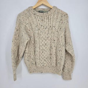 Könnte beinhalten: Ein beigefarbener Pullover mit Rundhalsausschnitt und einem komplexen Zopfstrickmuster mit vertikalen Säulen aus geflochtenen und rautenförmigen Texturen. Das Strickgewebe ist durchgehend mit kleinen schwarzen, blauen und roten Sprenkeln versehen.