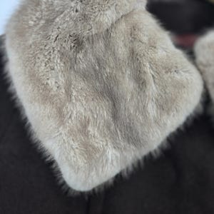 May include: Close-up of a light gray fur collar. The fur is dense and soft, with a slightly textured appearance. The collar is set against a dark brown background, creating a contrast that highlights the fur's color and texture. The collar appears to be part of a coat or jacket.