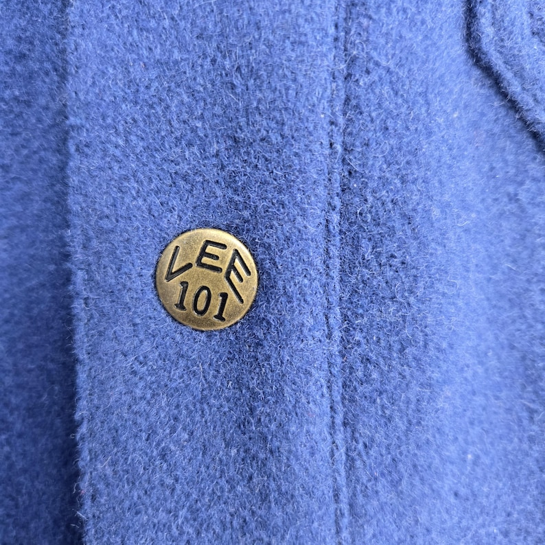 May include: Close-up of a blue fabric with a brass-colored button. The button is round with the text "LEE 101" embossed on it. The fabric has a textured appearance.