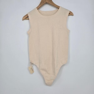May include: A sleeveless bodysuit in a solid cream color featuring a round neckline and a textured knit fabric. The garment is displayed on a wooden hanger and includes a small white product tag attached to the lower side seam near the hip area.