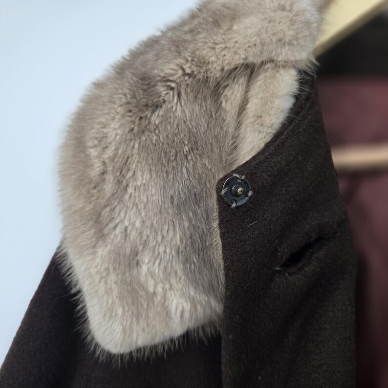 May include: Close-up of a dark brown coat with a light gray fur collar. The coat has a single button closure and a small slit detail. The fur collar adds a touch of luxury to this classic outerwear.