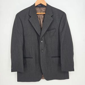 May include: A charcoal gray blazer featuring a subtle vertical texture, a notched lapel, and a two-button front closure. The garment includes a chest pocket, two lower flap pockets, and four buttons on each sleeve cuff, with a brown interior lining.
