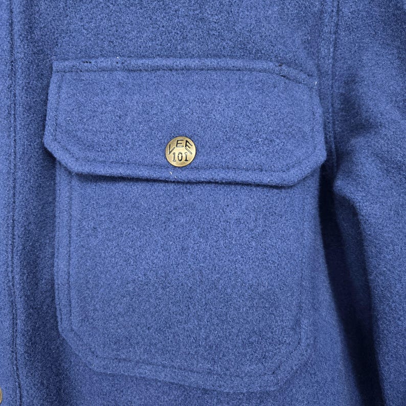 May include: Close-up of a blue jacket with a pocket and a gold-colored button. The button has the text "LEE 101" on it. The jacket is made of a textured fabric.