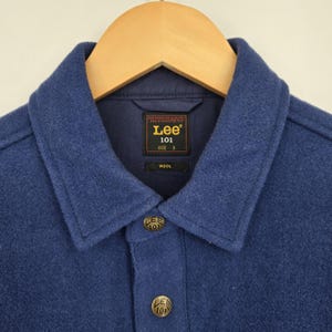 May include: Close-up of a blue wool jacket with a collar and button closure. The jacket features the "Lee 101" brand label and size "S". The buttons and label have the "Lee 101" logo. The jacket is hanging on a wooden hanger.