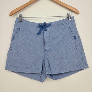 May include: Blue and white gingham shorts featuring a drawstring waist with a blue tie, two side pockets with blue accents, and a clean hemline. The shorts are displayed on a wooden hanger, showcasing a classic checkered pattern throughout.