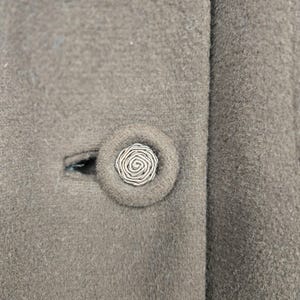 May include: Close-up of a taupe-colored coat with a decorative button. The button is round with a spiral design in the center, and is surrounded by a matching fabric. The coat has a classic, tailored look.