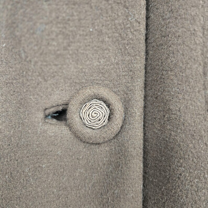 May include: Close-up of a taupe-colored coat with a decorative button. The button is round with a spiral design in the center, and is surrounded by a matching fabric. The coat has a classic, tailored look.