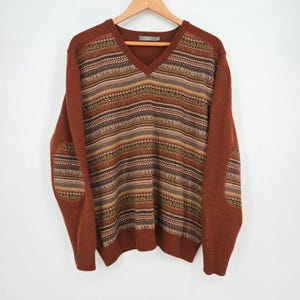 May include: A brown V-neck sweater featuring a horizontal patterned panel across the chest and back, with matching patterned elbow patches. The knit design includes stripes of geometric shapes and repeating text in shades of tan, olive, and dark brown.