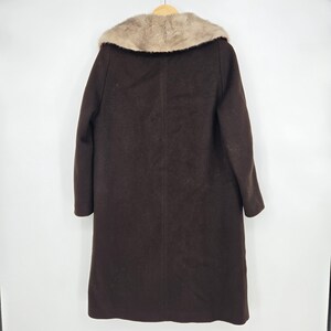 May include: A dark brown coat with a light brown fur collar hangs on a wooden hanger. The coat is long, reaching to the ankles, and has a simple, classic design. The fur collar adds a touch of luxury to the garment.