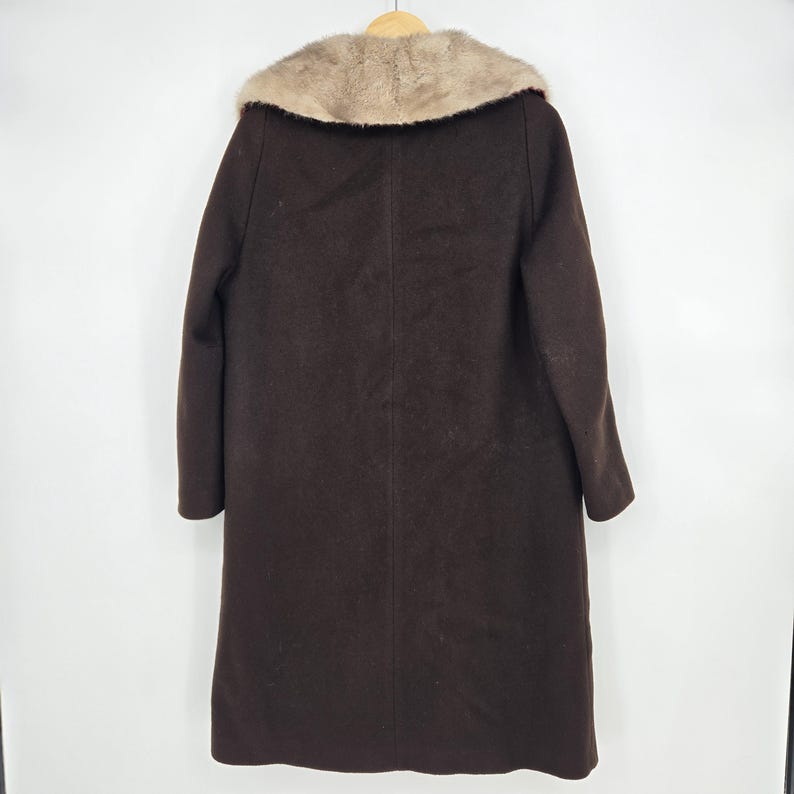 May include: A dark brown coat with a light brown fur collar hangs on a wooden hanger. The coat is long, reaching to the ankles, and has a simple, classic design. The fur collar adds a touch of luxury to the garment.
