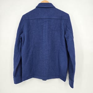 May include: A navy blue wool jacket with a button-down front and a collared neckline. The back of the jacket is visible, showing a simple, classic design. The jacket is hanging on a wooden hanger.