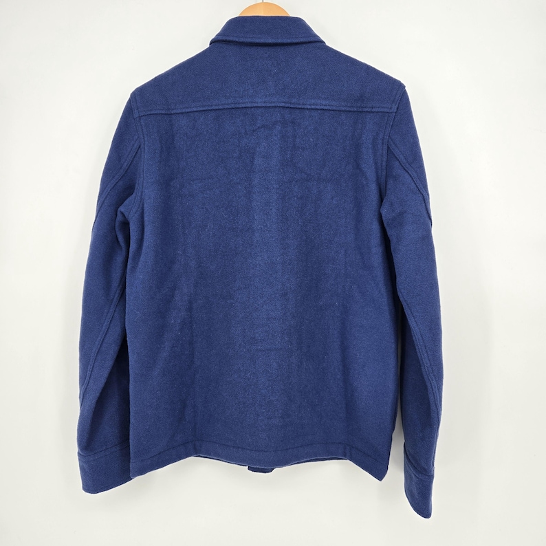 May include: A navy blue wool jacket with a button-down front and a collared neckline. The back of the jacket is visible, showing a simple, classic design. The jacket is hanging on a wooden hanger.