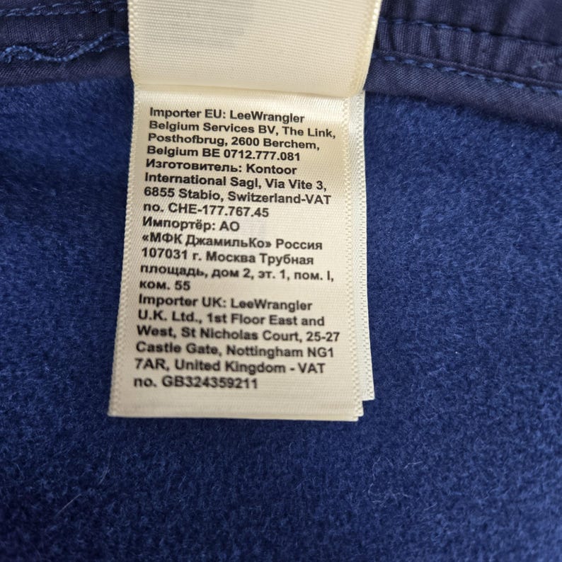May include: A close-up of a white tag with black text, attached to a blue fabric. The tag includes the text "Importer EU: LeeWrangler" and other details about the importer, including addresses and VAT numbers.
