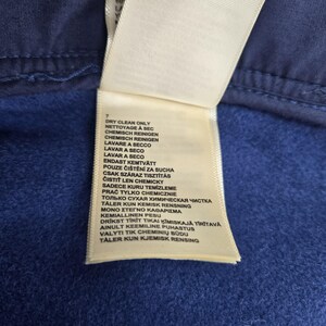 May include: Close-up of a white clothing tag with care instructions in multiple languages, including "Dry Clean Only." The tag is attached to a blue fabric, with a visible seam at the top.