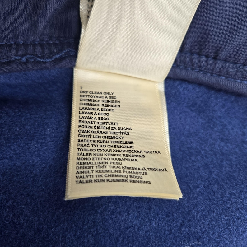 May include: Close-up of a white clothing tag with care instructions in multiple languages, including "Dry Clean Only." The tag is attached to a blue fabric, with a visible seam at the top.
