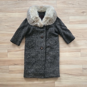 Puede incluir: Un abrigo vintage gris carbón con cuello de piel sintética. El abrigo tiene un aspecto texturizado, mangas tres cuartos y dos botones marrones en la parte delantera. El cuello es de color marrón claro y crema.