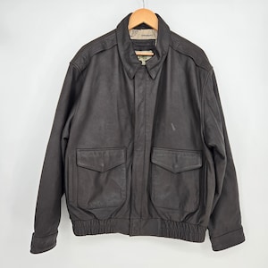 May include: A dark brown leather jacket with a collared neckline and two front pockets. The jacket has a ribbed hem and cuffs, and is hanging on a wooden hanger against a white background. The jacket appears to be vintage.