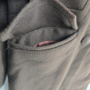 May include: Close-up of a brown wool coat pocket. The pocket is a deep brown color with a slightly textured surface. The interior lining of the pocket is a contrasting reddish-brown color. The coat appears to be made of a thick, warm material.