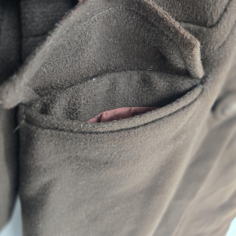 May include: Close-up of a brown wool coat pocket. The pocket is a deep brown color with a slightly textured surface. The interior lining of the pocket is a contrasting reddish-brown color. The coat appears to be made of a thick, warm material.