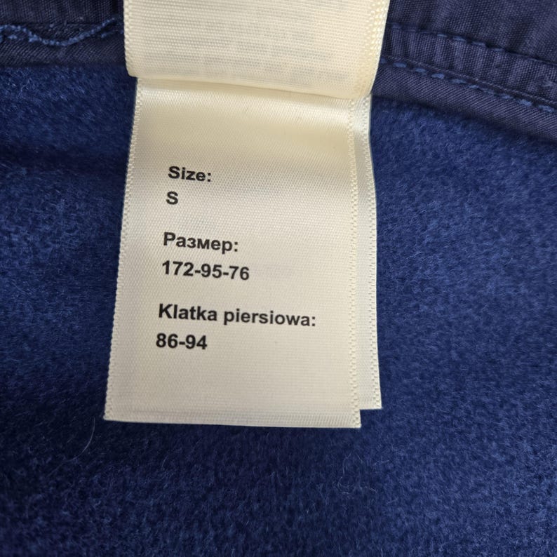 May include: A white clothing care label attached to a dark blue fleece garment. The label displays the text Size: S, followed by the Russian text for size 172-95-76, and the Polish text Klatka piersiowa: 86-94, indicating chest measurements.
